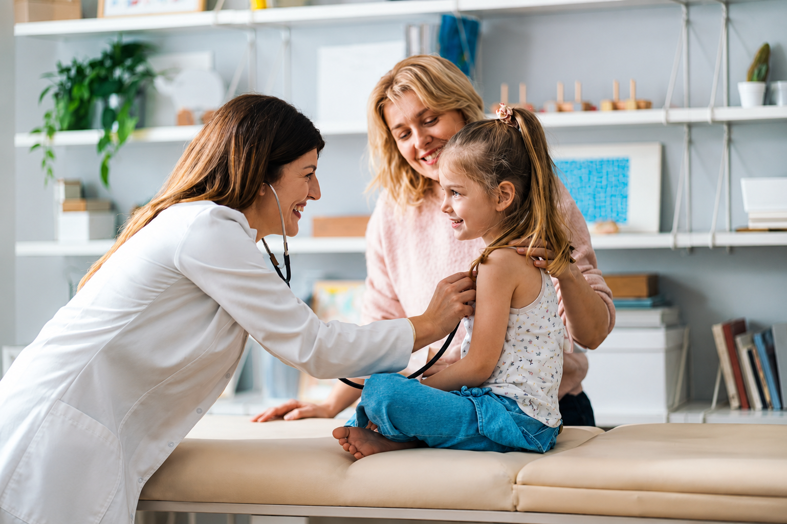 A pediatrician listens to a smiling young girl's heartbeat while her mother holds her